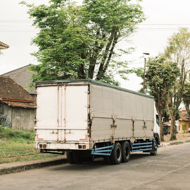 Home A heavy-duty cargo truck parked on a city street surrounded by lush greenery and buildings.